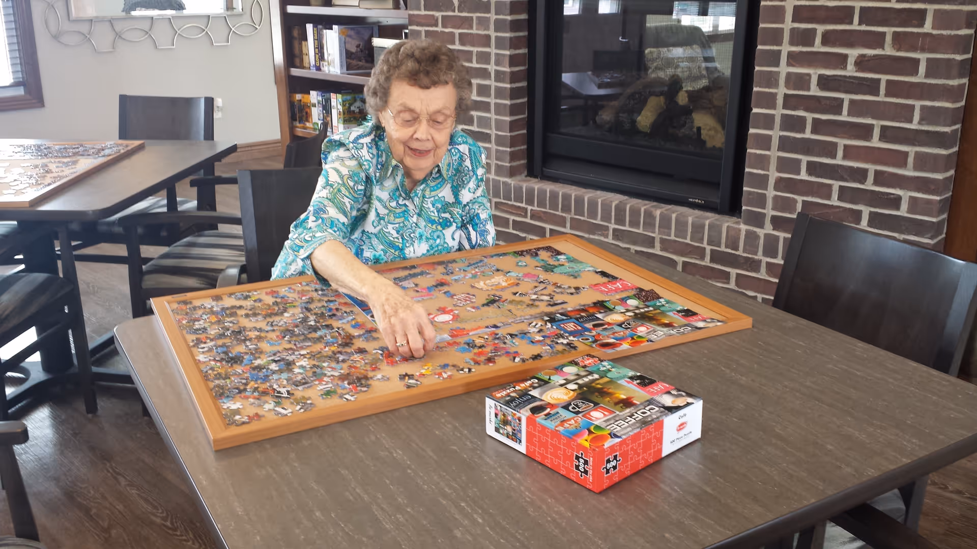 An elderly woman with glasses and curly hair is sitting at a table working on a large jigsaw puzzle in a room with a brick fireplace and bookshelves in the background.