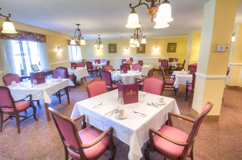A bright and neatly arranged dining room with multiple tables covered in white tablecloths, each set with cups, saucers, utensils, and a maroon menu card labeled Heatherwood Retirement Community. The room has pink cushioned chairs, yellow walls, framed artwork, and several ceiling light fixtures providing warm lighting.