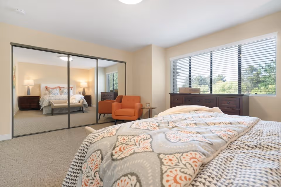 A bright bedroom with a large window featuring horizontal blinds. The bed is covered with a patterned comforter in shades of gray, white, and orange. There is a wooden dresser beneath the window and an orange armchair with a small side table next to it. A mirrored closet door reflects another view of the bed and bedside tables with lamps.
