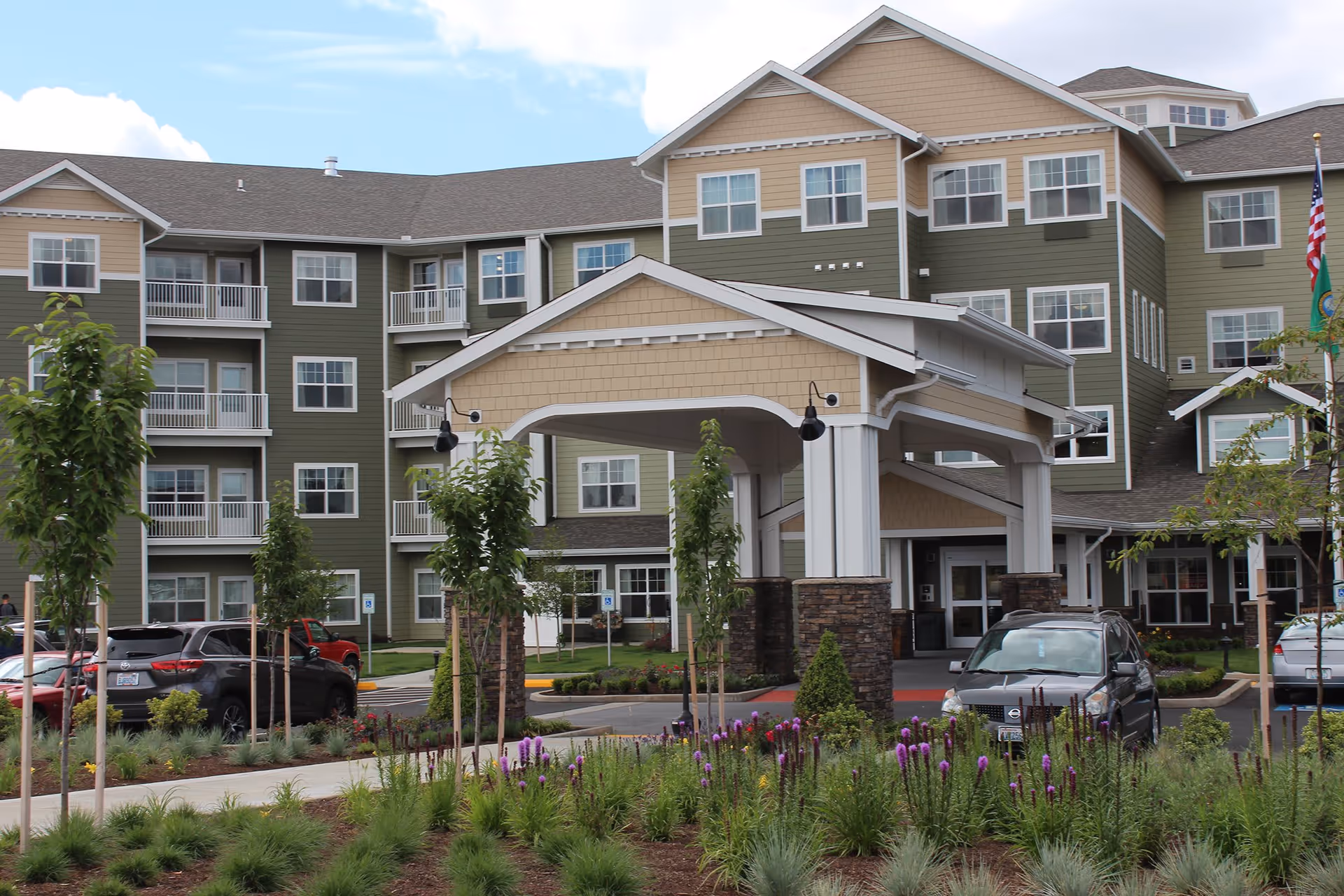 Exterior view of a multi-story senior living facility with green and beige siding, white balconies, and a covered entrance. There are several cars parked in front and landscaped greenery with small trees and purple flowers in the foreground.