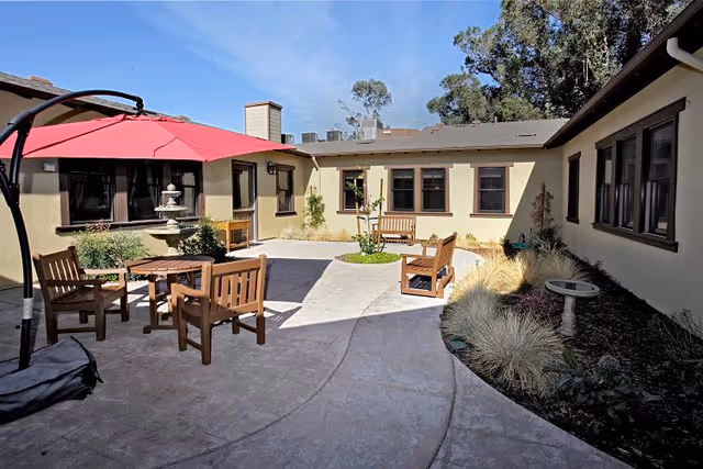 Outdoor courtyard area with wooden benches and a table under a large red umbrella. The courtyard is surrounded by a single-story building with multiple windows. There are plants and shrubs along the edges and a small birdbath on the right side.