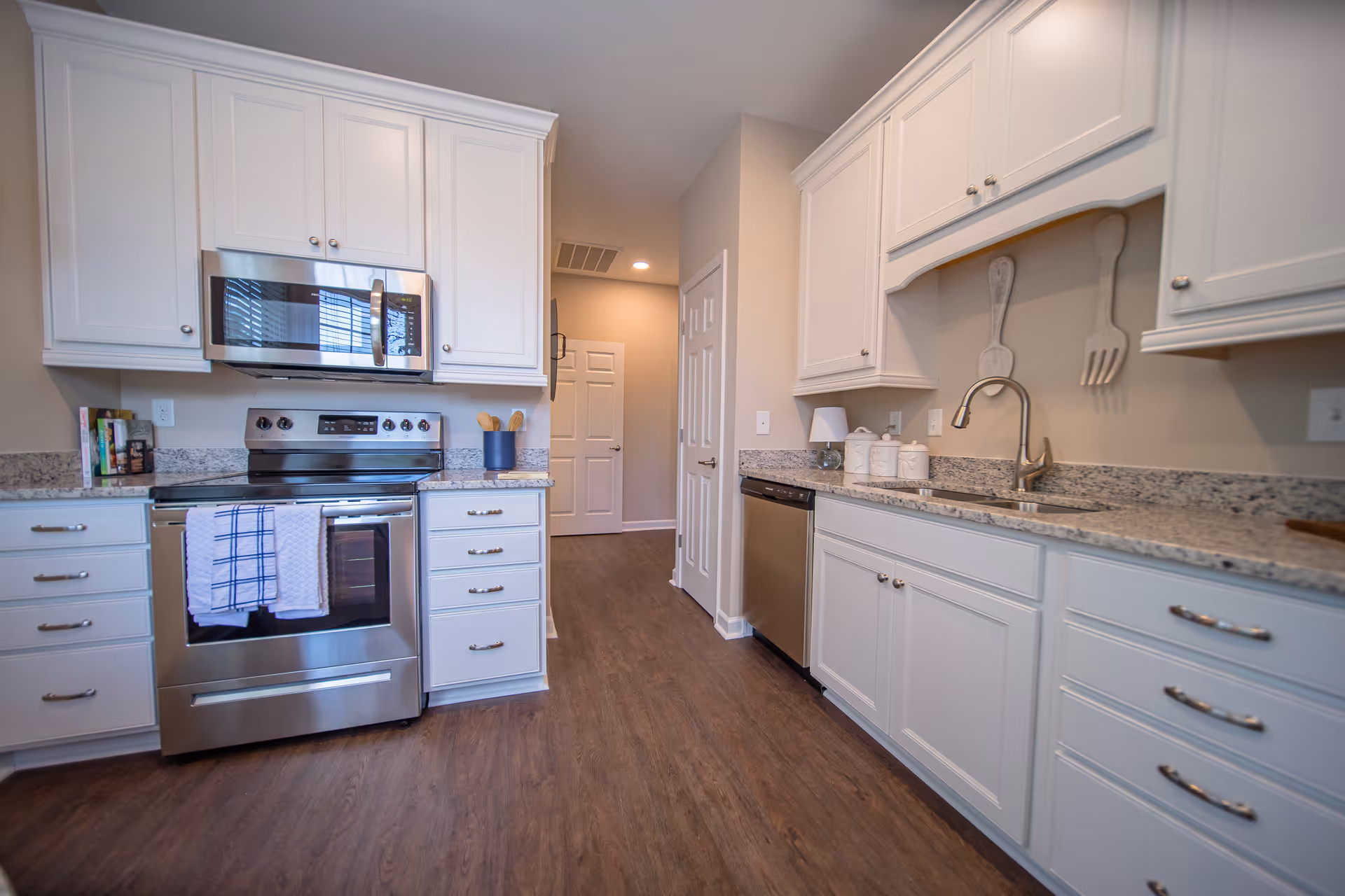 A modern kitchen with white cabinets, granite countertops, and stainless steel appliances including an oven and microwave. There are two kitchen towels hanging on the oven handle, a dishwasher next to the sink, and decorative oversized wooden fork and spoon hanging on the wall above the sink. The floor is wood, and there is a hallway visible in the background.