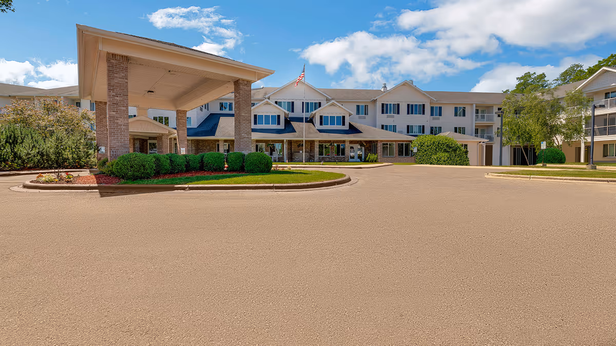 Exterior view of Middleton Crossing - Sky Active Living facility showing a large entrance canopy supported by brick columns, manicured bushes, and a flagpole with an American flag in front of a multi-story building under a partly cloudy blue sky.