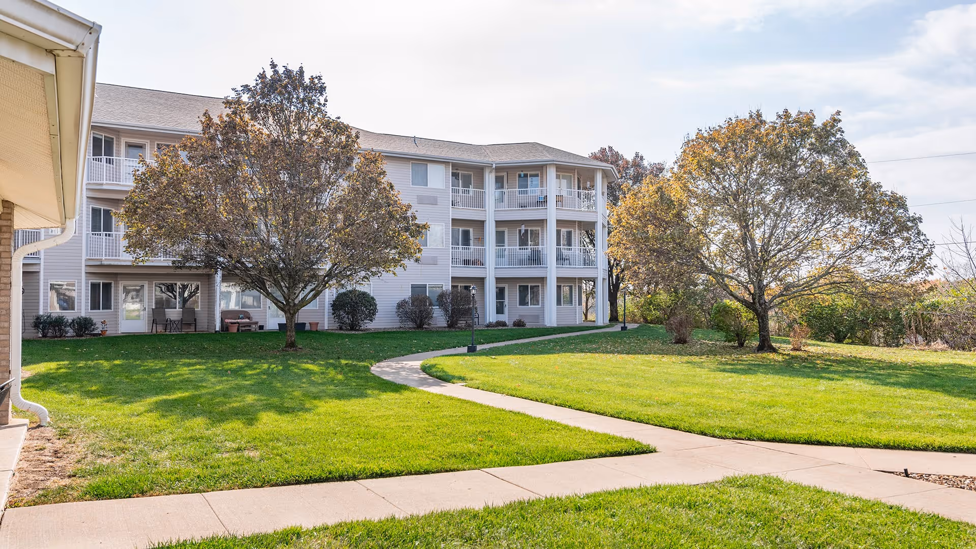 Outdoor view of a senior living facility with a three-story building in the background, surrounded by green grass, trees with autumn foliage, and a curved concrete walkway.