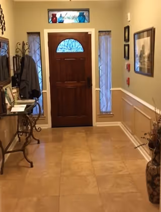 Interior view of an entryway with a wooden front door featuring a decorative glass window. The door is flanked by two narrow windows with frosted glass. On the left side, there is a small ornate table with framed photos and a coat hanging on a wall-mounted rack. The walls are painted light beige with white wainscoting, and there is a large decorative vase with dried plants on the right side.