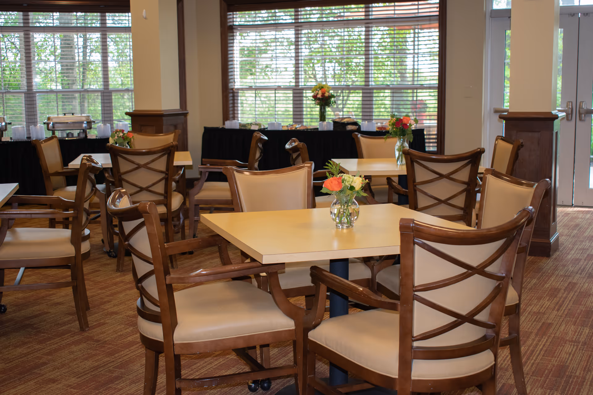 A dining room with several square tables surrounded by wooden chairs with beige cushions. Each table has a small glass vase with a floral arrangement. Large windows with wooden blinds let in natural light, and a buffet table with covered dishes and cups is visible in the background.
