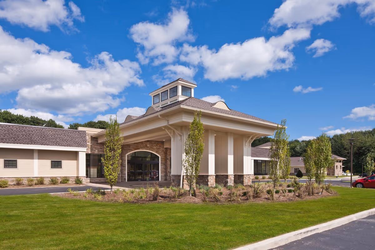 Exterior view of a senior living facility building with a covered entrance, stone and beige siding, surrounded by green grass, small trees, and a clear blue sky with scattered clouds.