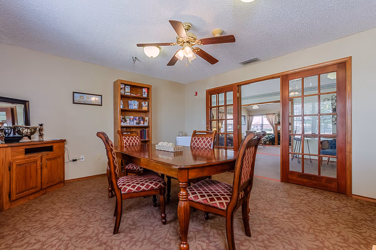 Wooden dining table with four upholstered chairs in a carpeted room with a ceiling fan and glass-paneled double doors.