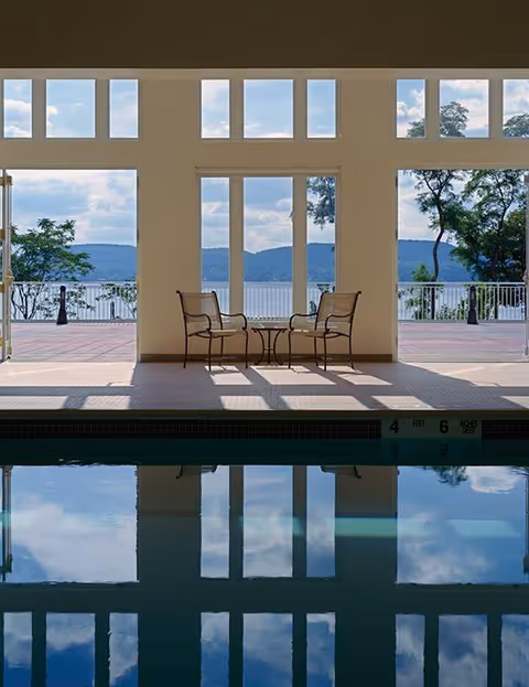 Indoor swimming pool area with large windows and glass doors opening to an outdoor patio with a view of trees and hills in the distance. Two metal chairs and a small round table are positioned near the windows, with reflections visible on the pool water.