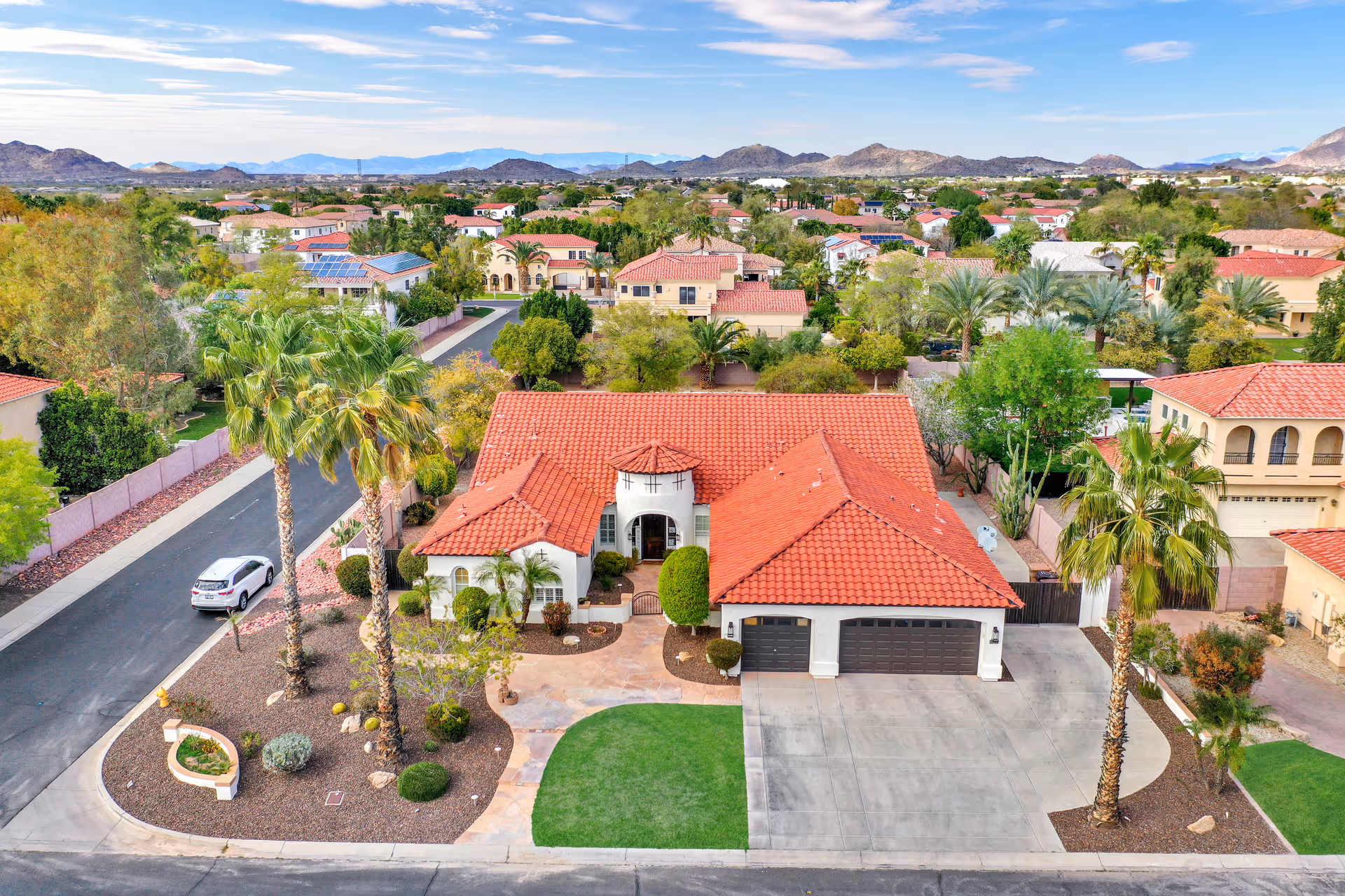 Aerial view of a single-story house with a red tile roof, a three-car garage, and a landscaped front yard featuring palm trees and shrubs. The house is located in a suburban neighborhood with similar houses and mountains visible in the background under a partly cloudy sky.