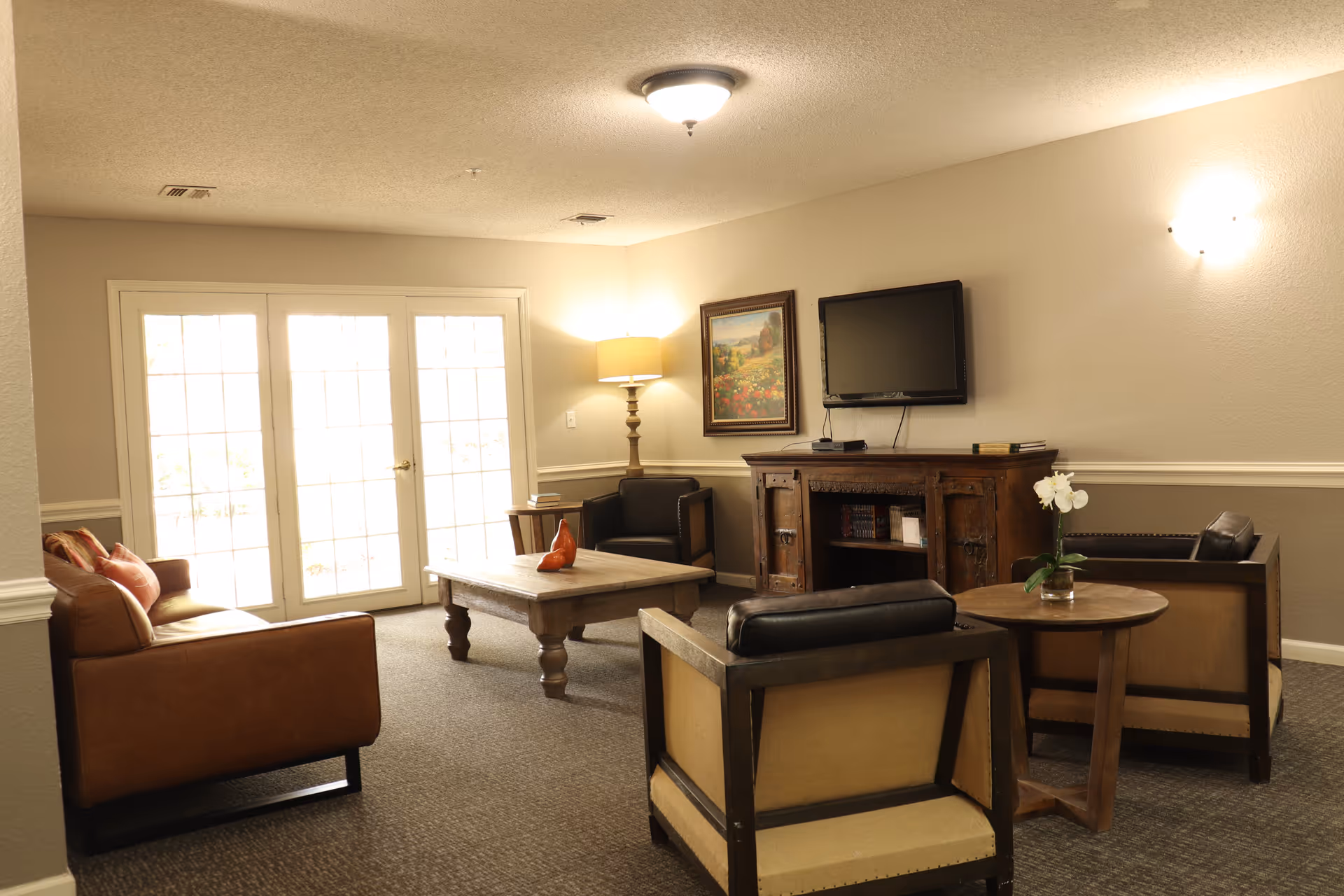 A cozy living room with a brown leather sofa, two armchairs, a wooden coffee table, and a side table with a small plant. There is a wooden cabinet with a flat-screen TV mounted above it, a floor lamp in the corner, and a framed painting on the wall. Large glass doors allow natural light to fill the room.