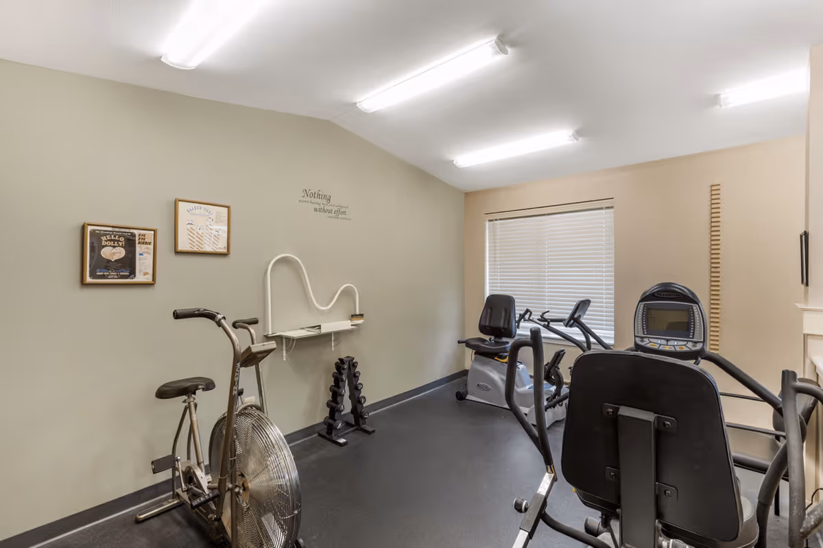 A small exercise room with stationary bikes, a recumbent bike, and a set of dumbbells on a rack against a beige wall. The room has fluorescent ceiling lights, a window with closed blinds, and framed pictures on the wall.
