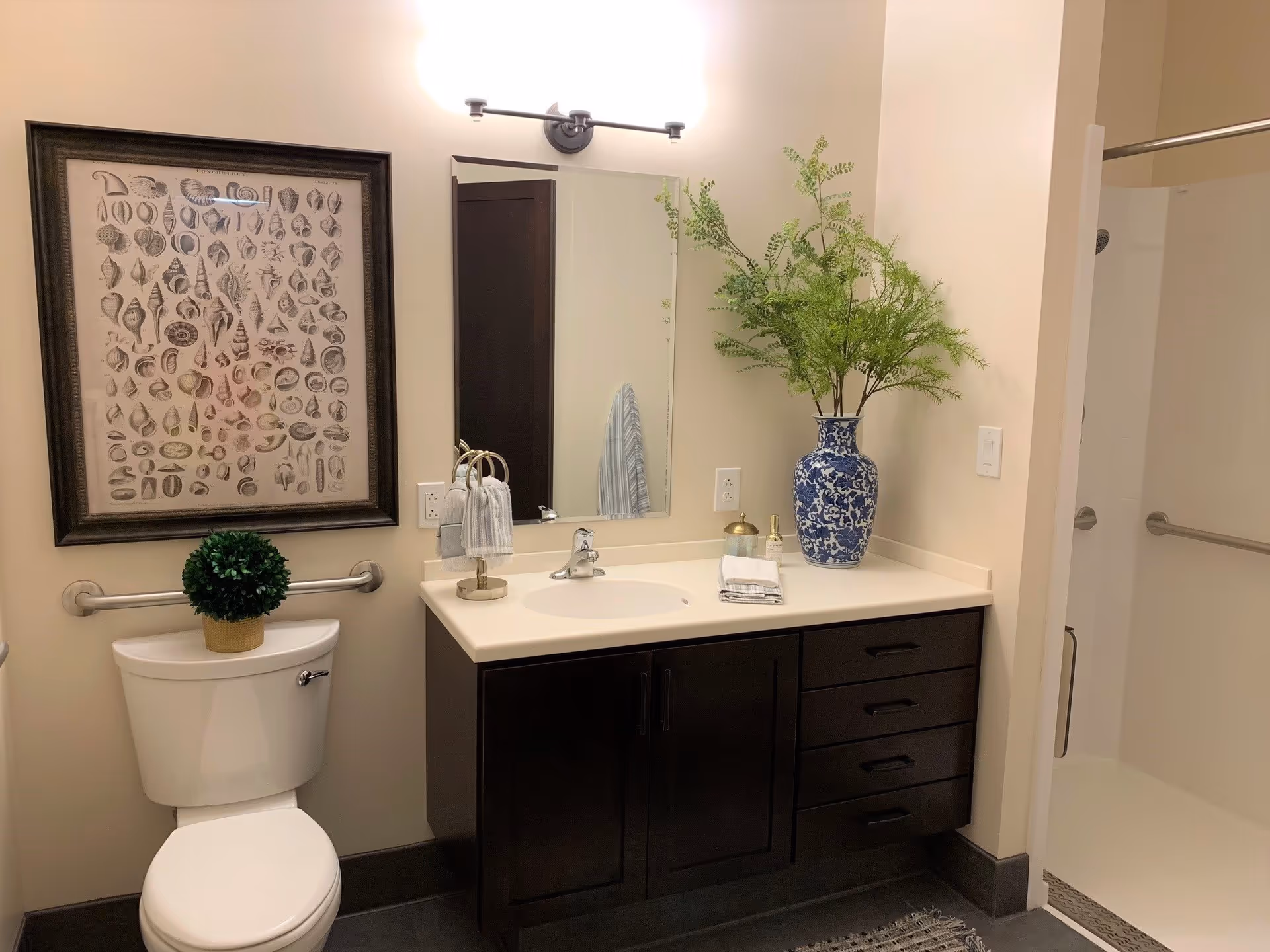 A clean and modern bathroom featuring a white toilet with a small green plant on top, a dark wood vanity with a white countertop and sink, a large mirror above the sink, a blue and white vase with green foliage on the countertop, a towel holder with hand towels, a framed artwork of seashells on the wall, and a walk-in shower with a grab bar.