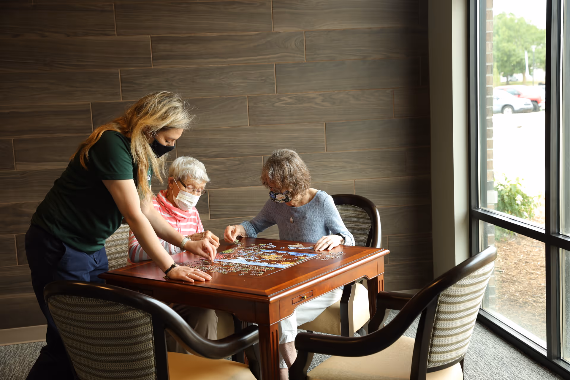 Three women wearing face masks are sitting and standing around a wooden table working on a jigsaw puzzle in a well-lit room with a large window and wood-paneled wall.