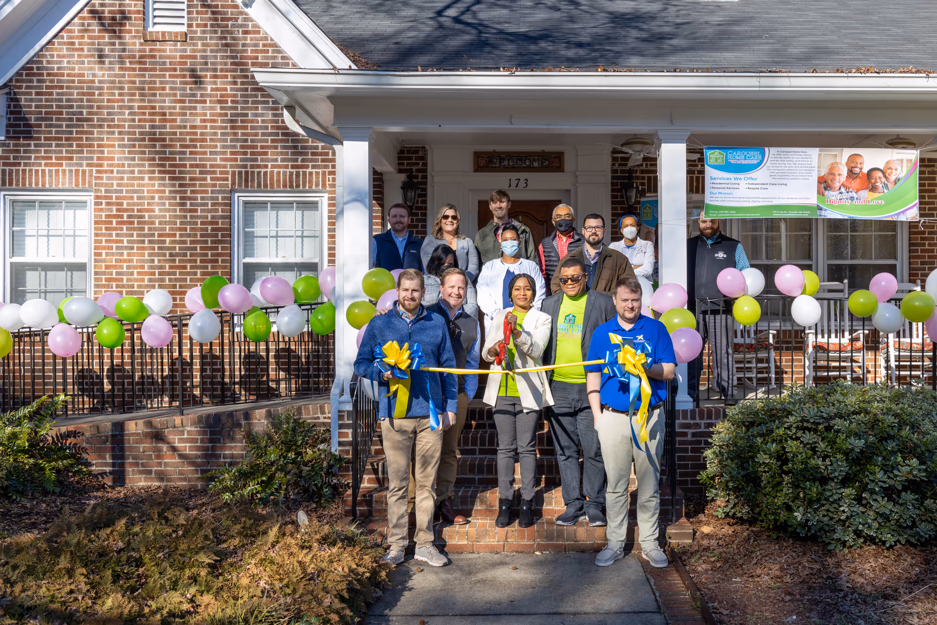 A group of people standing in front of a brick building decorated with pink, green, and white balloons. The group is gathered for a ribbon-cutting ceremony, with two men holding large scissors cutting a yellow ribbon. A banner for Carousel Home Care is displayed on the building's porch.
