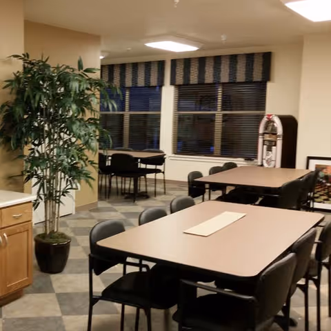 Interior view of a communal dining area with multiple rectangular tables and black chairs arranged around them. There is a large potted plant on the left side near a wooden cabinet. In the background, there are windows with blinds and a jukebox near the wall.
