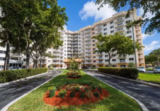 Exterior view of a multi-story senior living facility with balconies, surrounded by trees and landscaped gardens under a blue sky with some clouds.