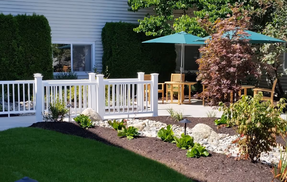 Outdoor patio area with wooden chairs and tables under green umbrellas, surrounded by landscaped garden beds with plants, rocks, and mulch. A white railing fence separates the patio from a grassy lawn. The exterior of a building with windows and green bushes is visible in the background.