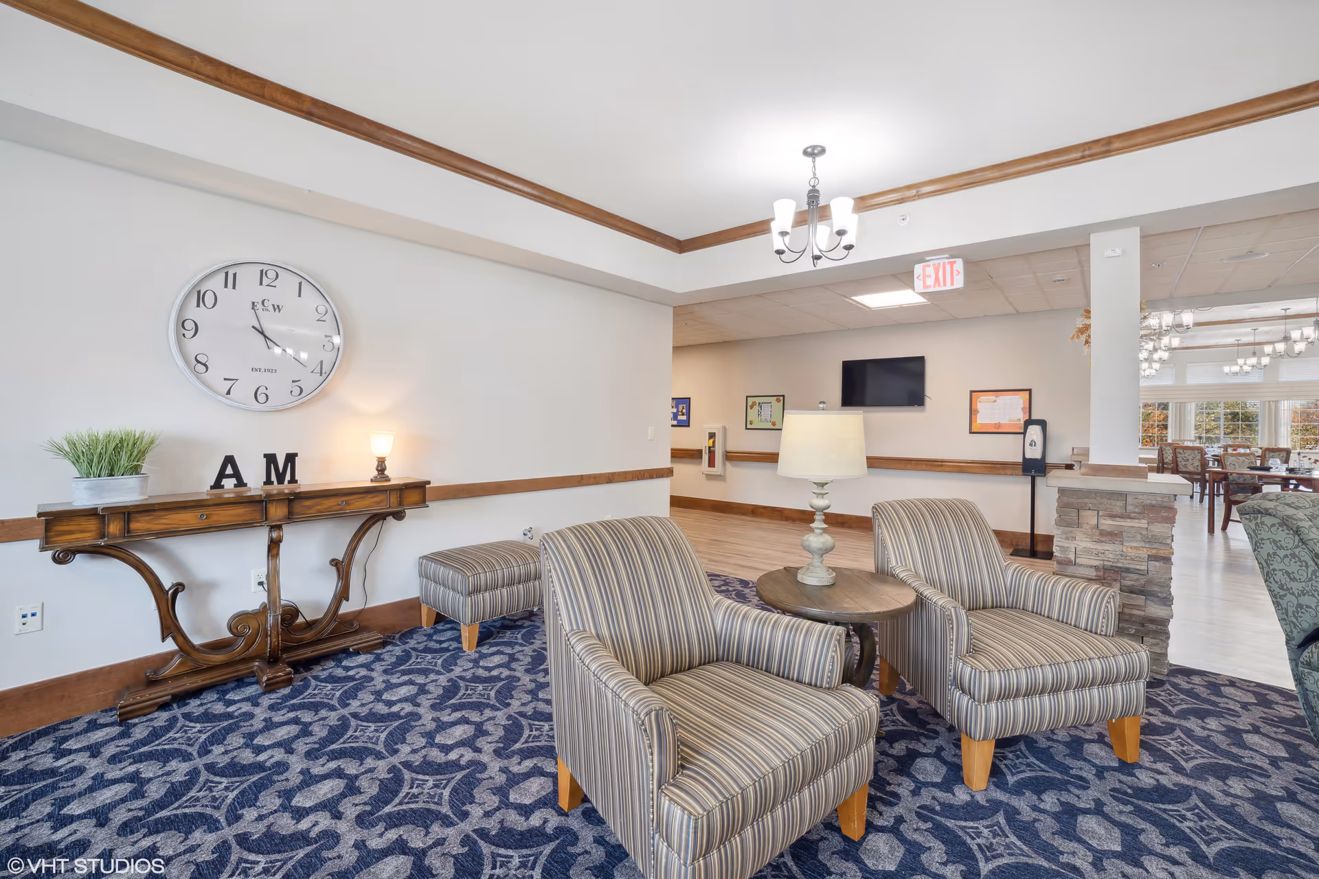 A cozy seating area in a senior living facility with two striped armchairs around a small round wooden table with a lamp. Behind the chairs is a wooden console table with a small lamp, a plant, and decorative letters 'A' and 'M'. A large wall clock hangs above the console table. The room has a patterned blue carpet, white walls with wooden trim, and a chandelier on the ceiling. In the background, there is a hallway with an exit sign and a dining area with tables and chairs near large windows.