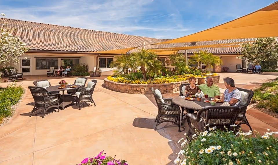Outdoor courtyard area at Silver Creek Inn Memory Care with several round tables and cushioned chairs. Three elderly people are seated at one table enjoying a meal. The courtyard features a central raised flower bed with palm plants and yellow flowers, shaded by large tan fabric canopies. The building with a tiled roof surrounds the courtyard, and additional seating is visible near the building.