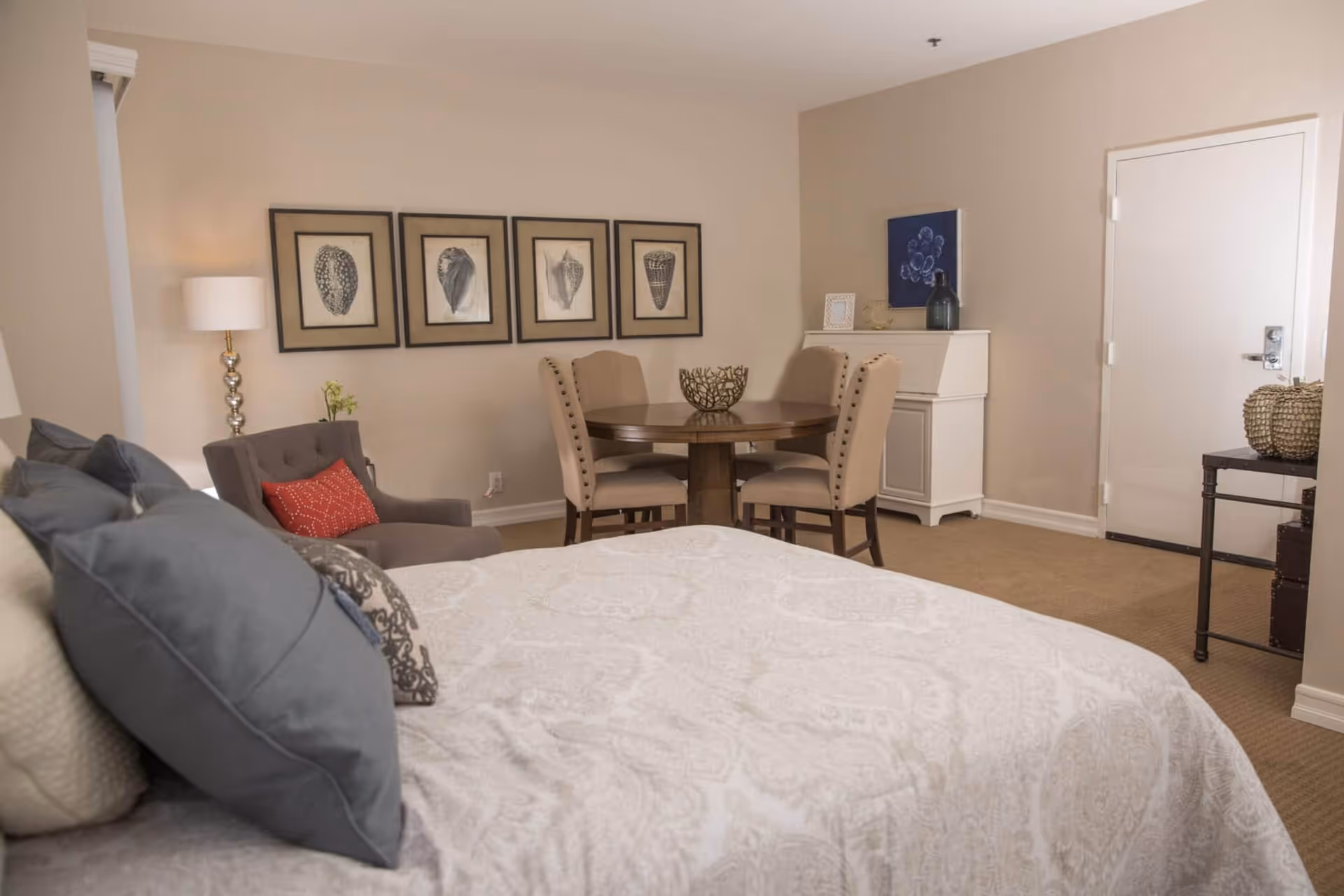 A furnished studio-style bedroom with a bed in the foreground, an armchair and lamp to the left, and a small dining table and cabinet against a beige wall.