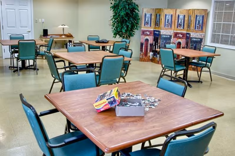 A room with several square wooden tables surrounded by teal cushioned chairs. On the nearest table, there is an open puzzle box with puzzle pieces scattered around it. In the background, there is a decorative folding screen with artwork and a tall green plant near the wall. The room has light-colored walls and a window with blinds.