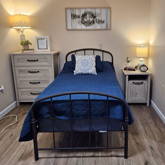 A neatly made single bed with a dark metal frame and a blue quilted bedspread, featuring a decorative pillow in the center. On the left side of the bed is a wooden dresser with four drawers, a table lamp, a small plant, and a framed picture. On the right side is a wooden nightstand with a drawer and cabinet, topped with a table lamp, a small plant, a book, and an alarm clock. Above the bed hangs a wooden sign that reads 'Bless All Who Gather Here.' The room has wood flooring and beige walls.