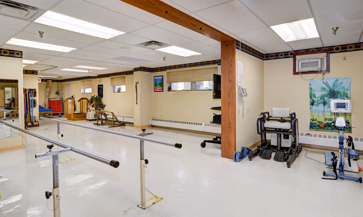 Bright physical therapy room with parallel bars, exercise machines, treatment equipment, and a wooden support column.