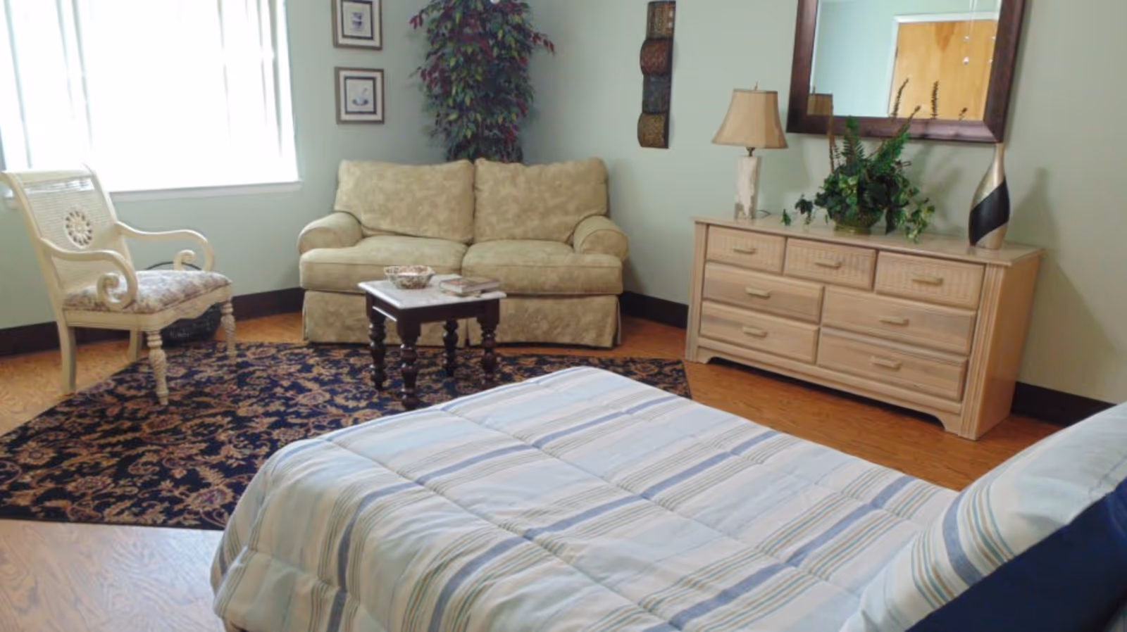 A cozy bedroom with a bed covered in a striped comforter in the foreground, a beige patterned loveseat, a wooden chair with a floral cushion, a small wooden coffee table, a dresser with a lamp, a plant, and a decorative vase, a large mirror on the wall, and a window letting in natural light.