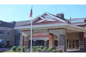 Exterior view of a senior living facility building with a covered entrance supported by stone pillars, an American flag on a flagpole in front, and well-maintained landscaping.