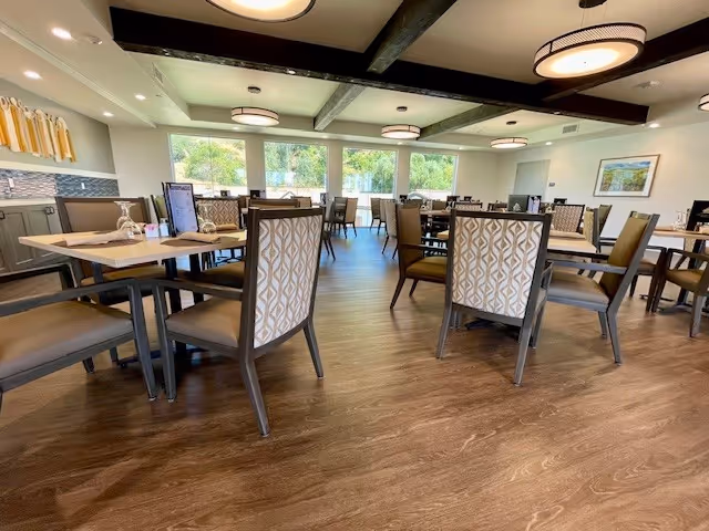A spacious dining room with multiple tables and chairs arranged neatly. The room has large windows letting in natural light, wooden flooring, and ceiling lights with circular fixtures. The chairs have patterned backs and cushioned seats, and the tables are set with menus and glassware.