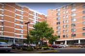 Exterior view of a multi-story brick senior living building with a landscaped entrance, parked cars, and trees.