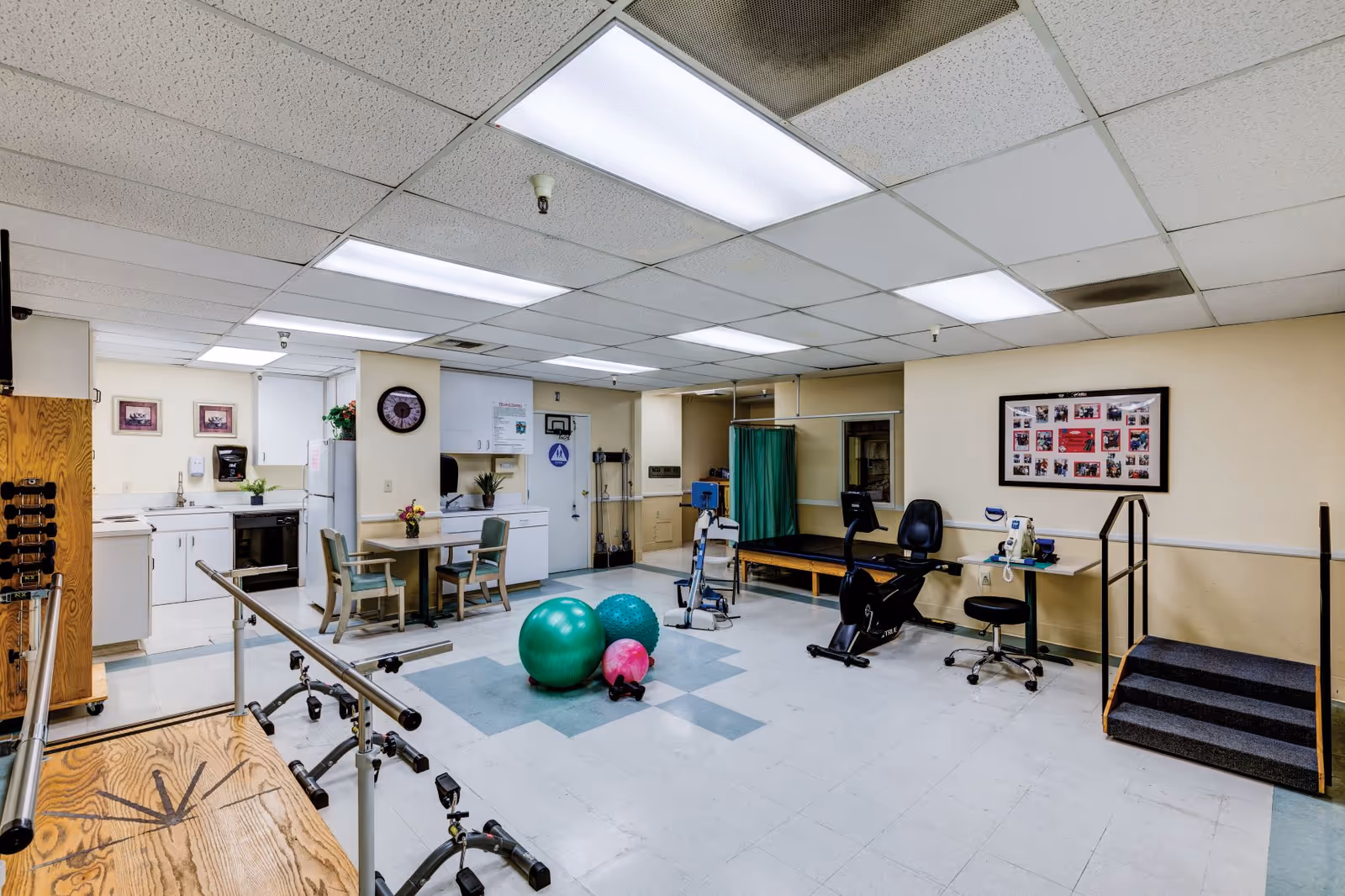 Therapy room with parallel bars, exercise balls, a stationary bike, treatment table, kitchenette, and other rehab equipment.