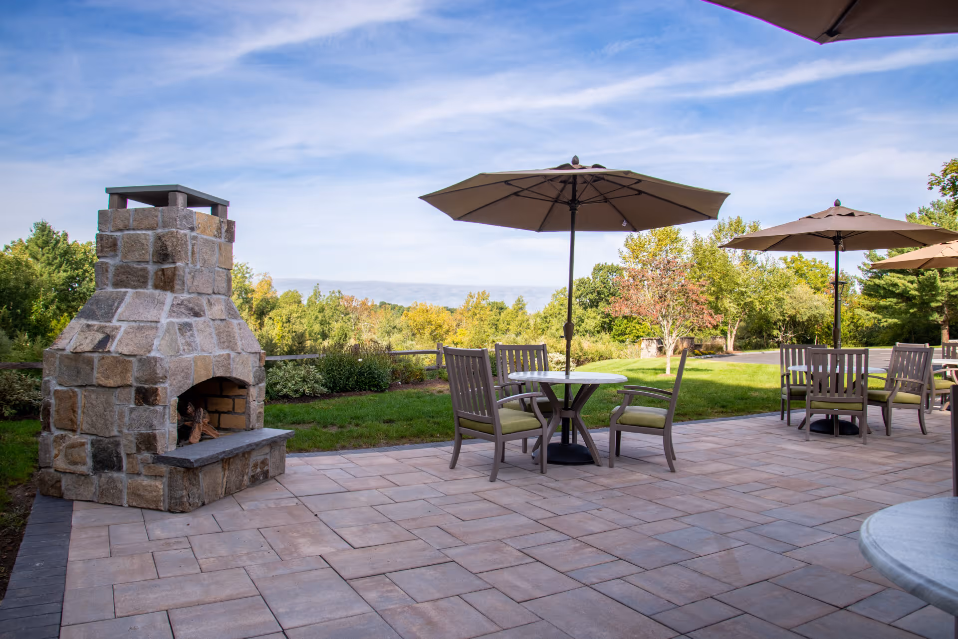 Outdoor patio with a stone fireplace, round tables, chairs and umbrellas overlooking a landscaped lawn and trees.
