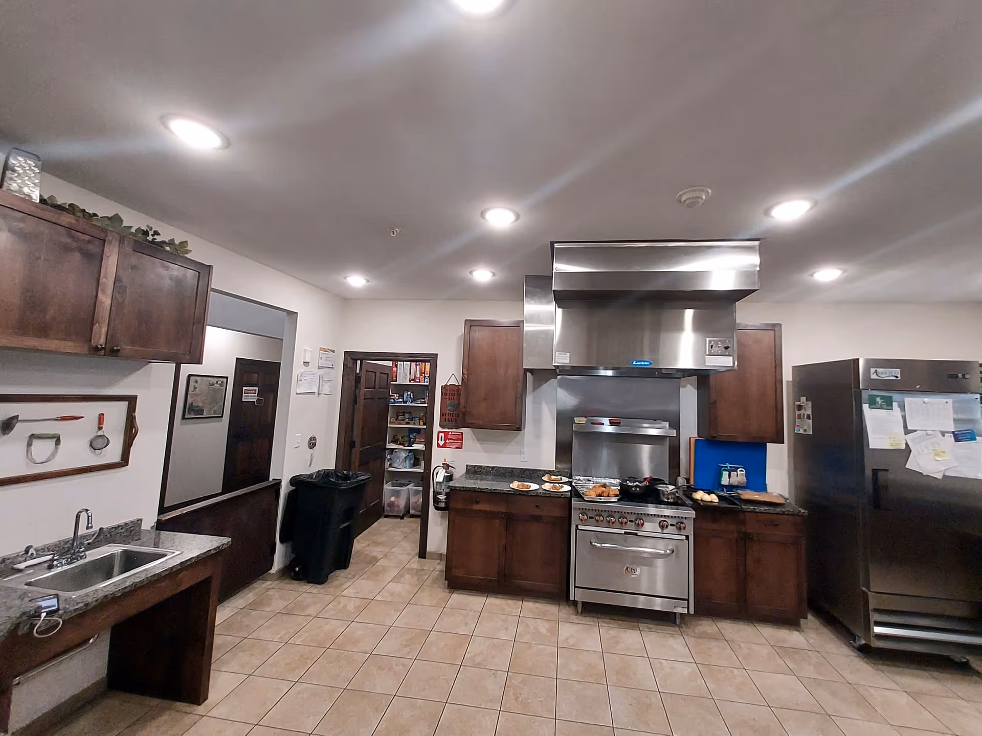 Interior view of a commercial kitchen with stainless steel stove and hood, wooden cabinets, a large refrigerator with papers attached, a double sink, and tiled floor. Various kitchen items and food are placed on the countertops.