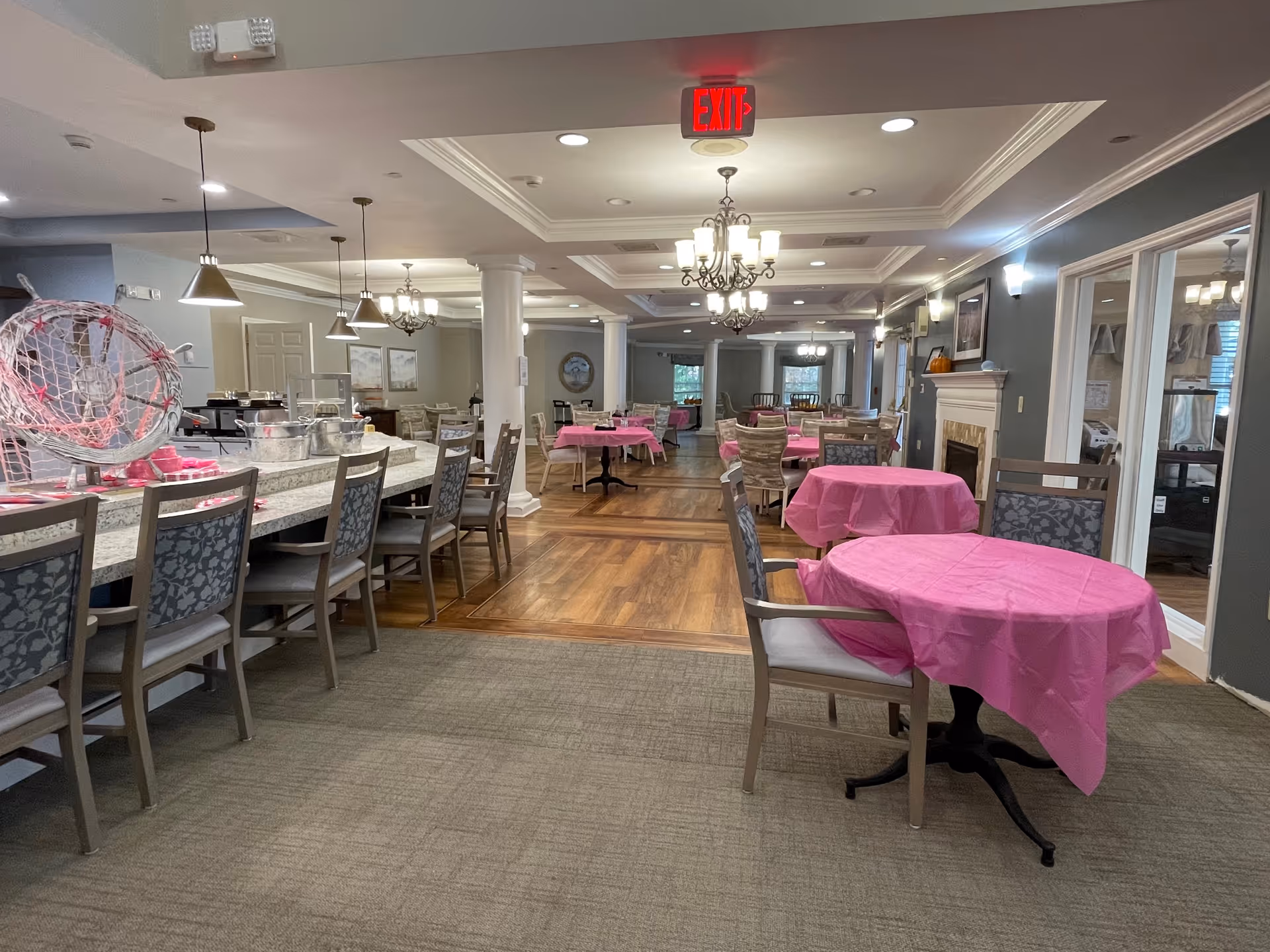 A spacious dining room in a senior living facility with multiple tables covered in pink tablecloths and surrounded by chairs. There is a long counter with chairs on one side, pendant lights hanging above, and chandeliers on the ceiling. The room features a fireplace on the right wall and large windows allowing natural light.