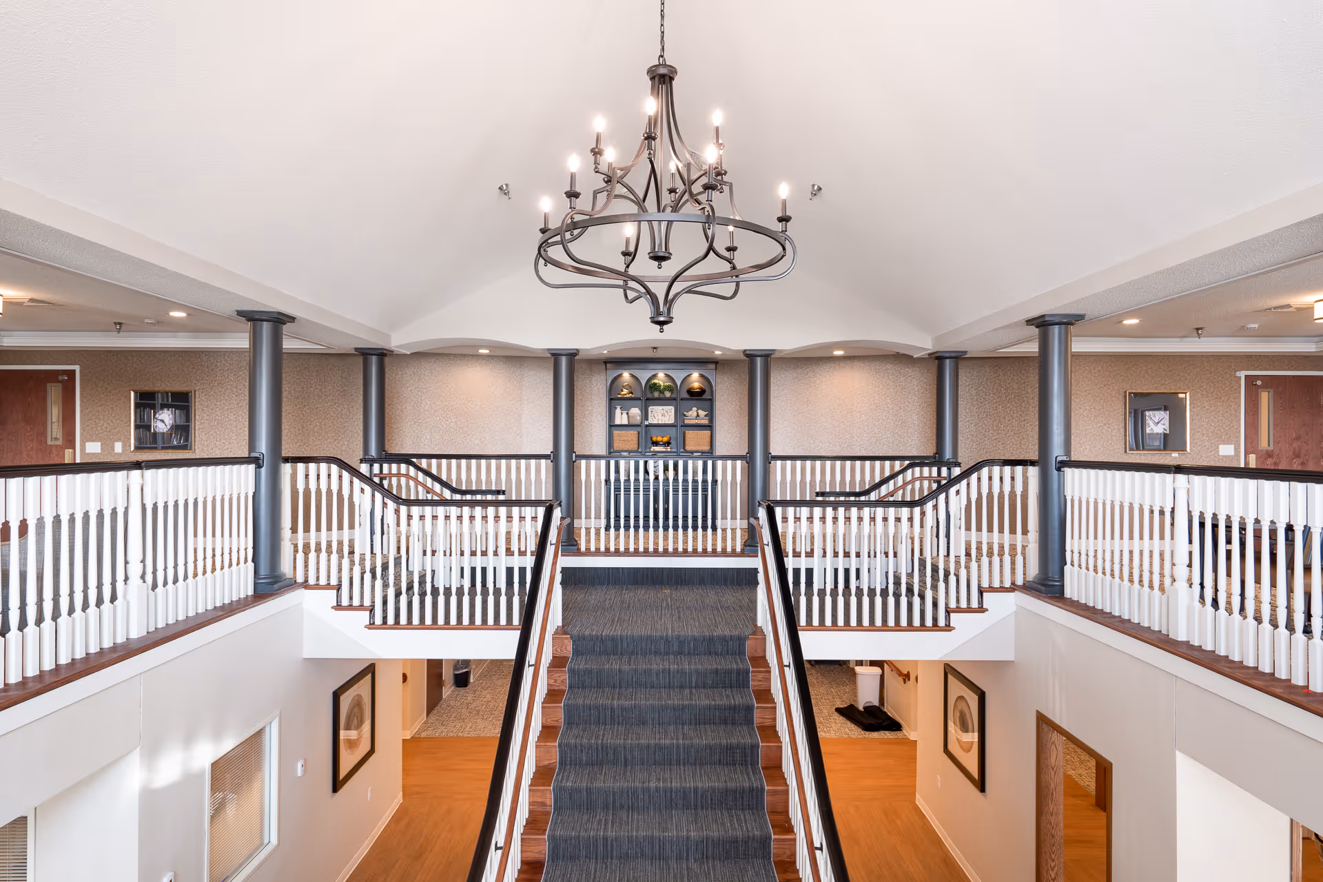 Interior view of a senior living facility featuring a wide staircase with a dark carpet runner leading to an upper level with white railings and black posts. A large decorative chandelier hangs from the ceiling above the staircase. The upper level has a built-in shelving unit with decorative items and two doors on either side. The walls are light-colored with subtle patterns, and the floor on the lower level is wood with framed artwork on the walls.