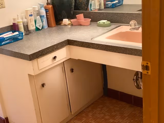 A bathroom countertop with various personal care products including lotions, sprays, and soaps. The countertop is gray with a pink sink embedded. Below the countertop are white cabinets and drawers. A mirror is visible above the counter reflecting some of the items.
