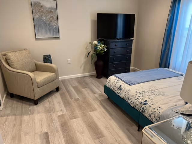 A furnished bedroom with a bed, armchair, dresser topped by a TV, and a vase of flowers by the wall.