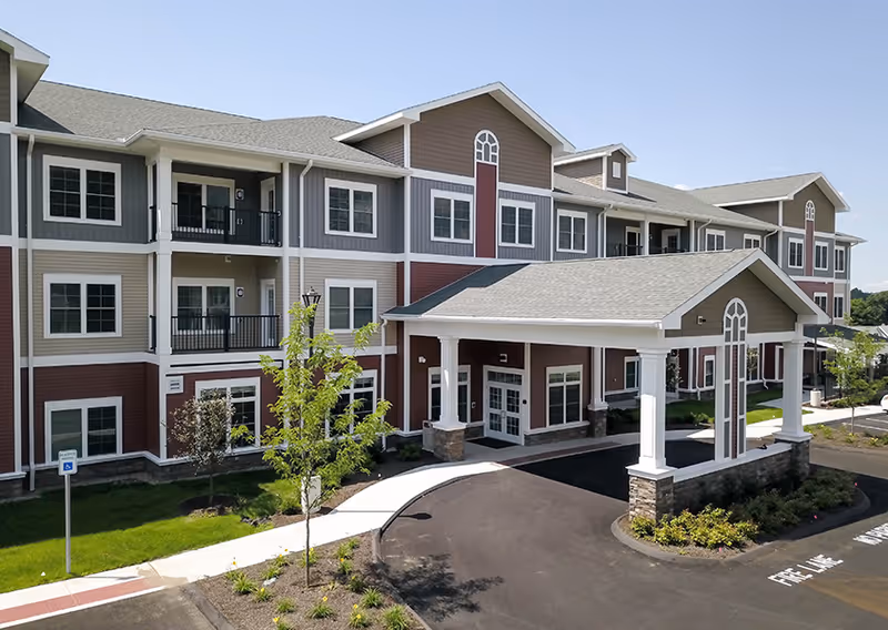 Front entrance and porte-cochère of a three-story senior living building with balconies, landscaped grounds, and a paved driveway.