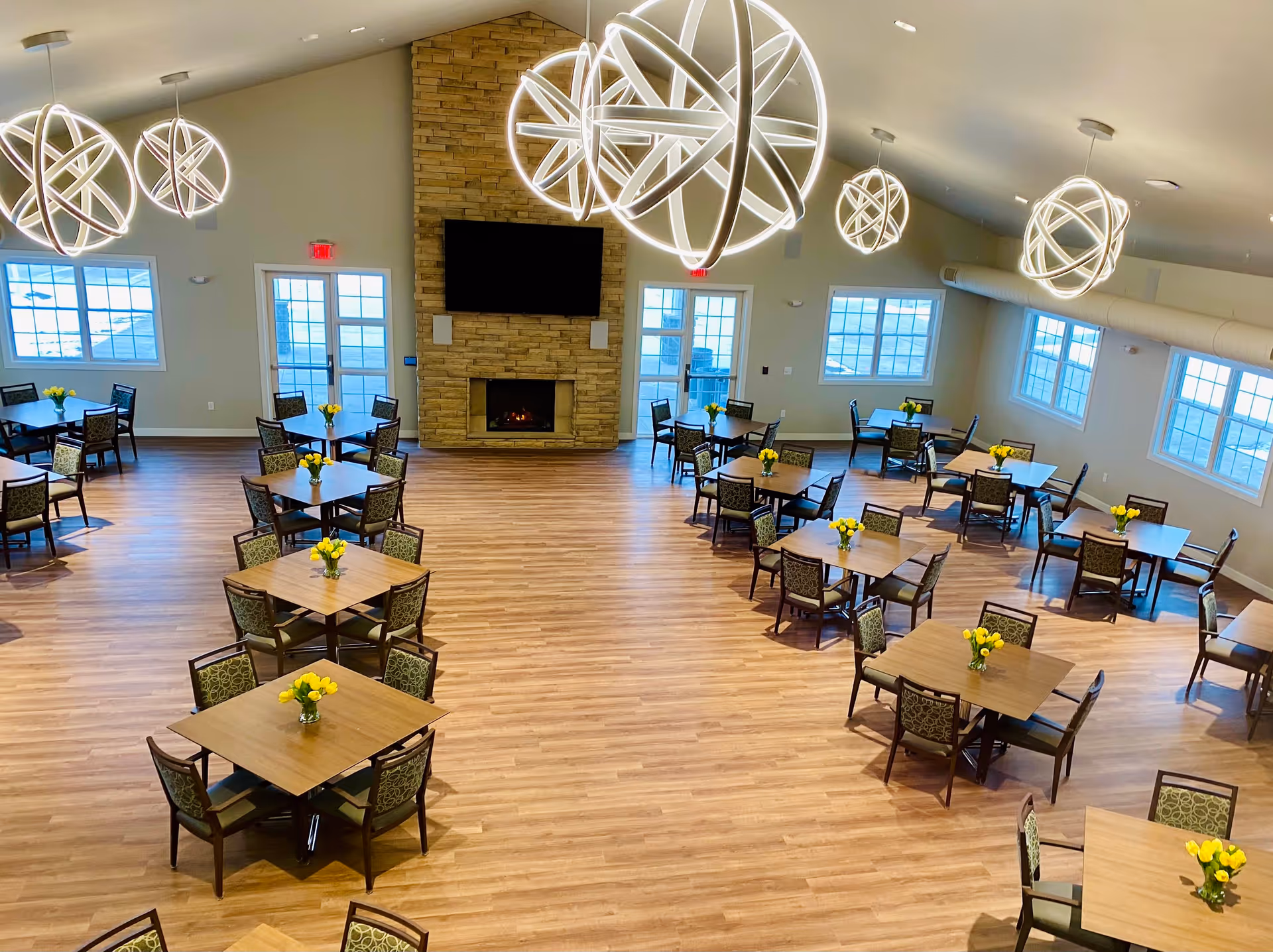 Spacious dining room with multiple square wooden tables each surrounded by four chairs. Each table has a small vase with yellow flowers. The room features large windows, modern spherical pendant lights hanging from the ceiling, and a stone fireplace with a mounted flat-screen TV above it.