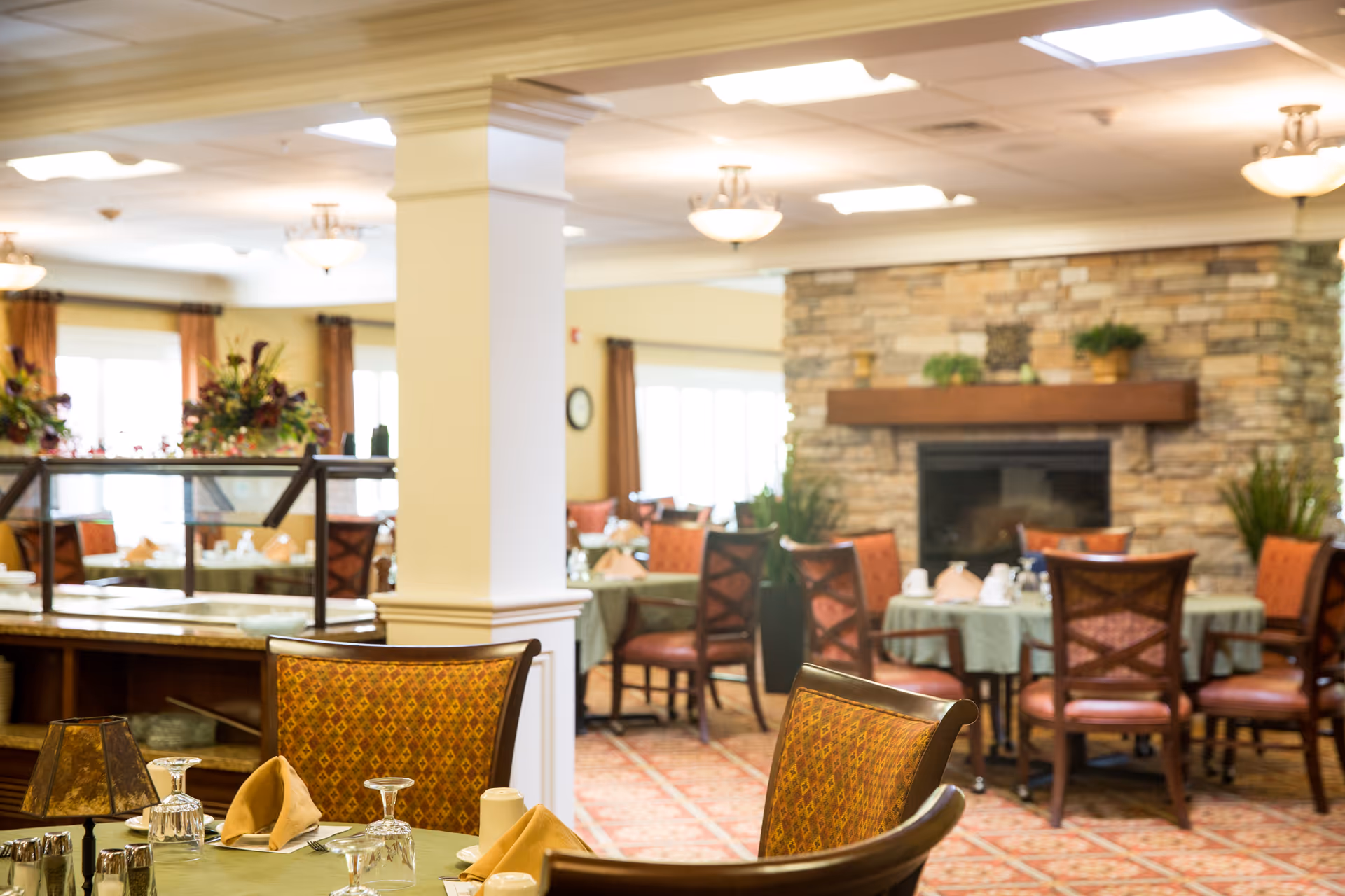 A cozy dining room in a retirement community featuring tables set with green tablecloths, folded napkins, glassware, and silverware. The room has warm lighting, a stone fireplace with plants on the mantel, and large windows with curtains allowing natural light to enter.