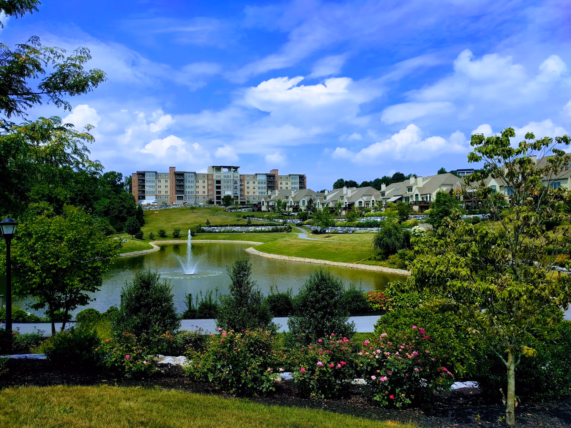 Scenic view of a senior living community with a central pond and fountain, landscaped gardens, and multi-story residential buildings under a blue sky.