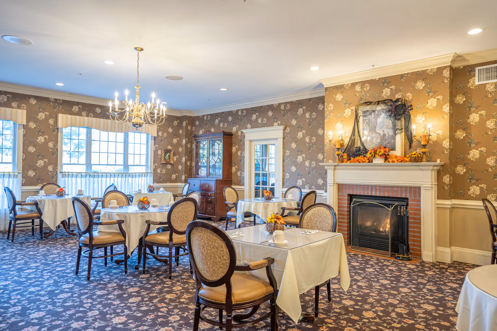 A cozy dining room in an assisted living community with round tables covered in white tablecloths, each set with cups and small floral centerpieces. The room features floral wallpaper, a chandelier, large windows with curtains, a wooden cabinet, and a fireplace decorated with autumn-themed decor.