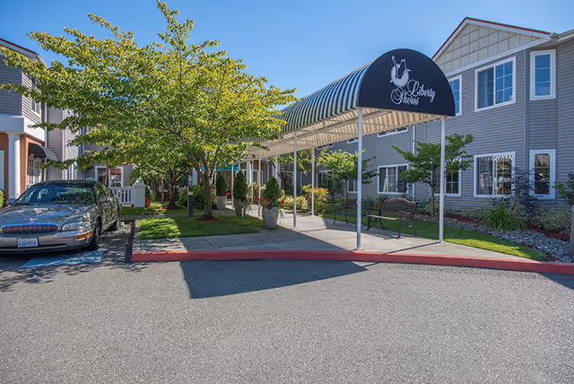 Exterior view of Liberty Shores Assisted Living facility showing a covered walkway with a black awning displaying the facility's name and logo. There are trees, benches, and a parked car near the entrance on a sunny day.