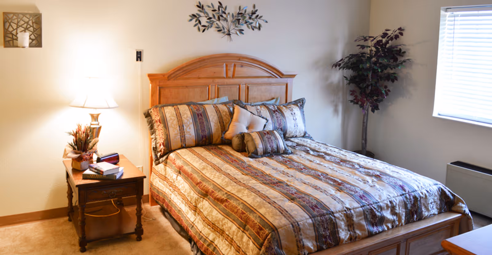 A cozy bedroom with a wooden bed frame and a patterned bedspread with multiple pillows. There is a wooden nightstand with a lamp, books, and a small plant. A decorative metal wall art piece is above the bed, and a potted plant is in the corner near a window with blinds.