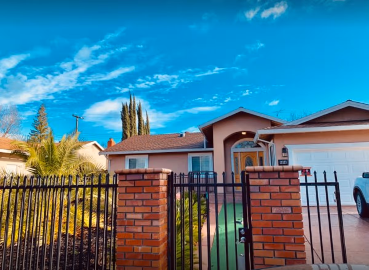 Front exterior of a single-story house with a gated brick fence, driveway and garage under a bright blue sky.