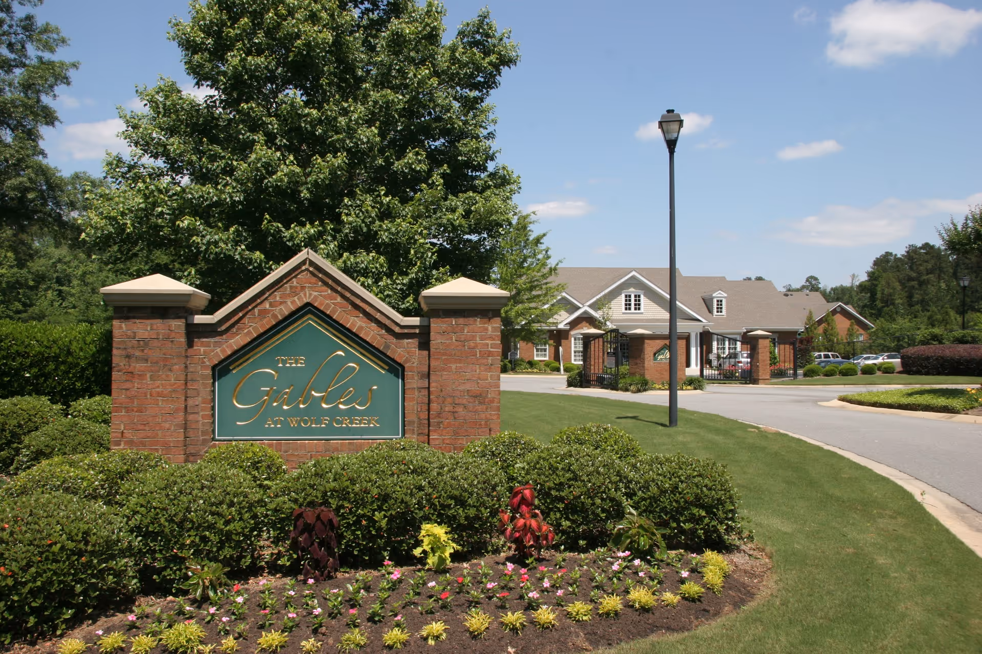 Entrance sign for The Gables at Wolf Creek, a senior living facility, surrounded by well-maintained bushes and flowers with a large tree behind it. In the background, there is a paved driveway leading to a building with a gray roof and brick pillars, under a clear blue sky.