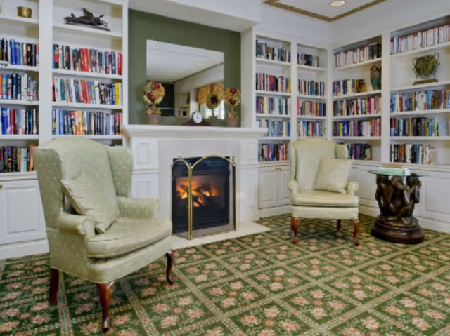 Cozy interior room with two green upholstered armchairs facing a lit fireplace surrounded by built-in white bookshelves filled with books. A decorative mirror and floral arrangements are on the mantel, and a small round wooden table is beside one chair. The floor is covered with a patterned green carpet.