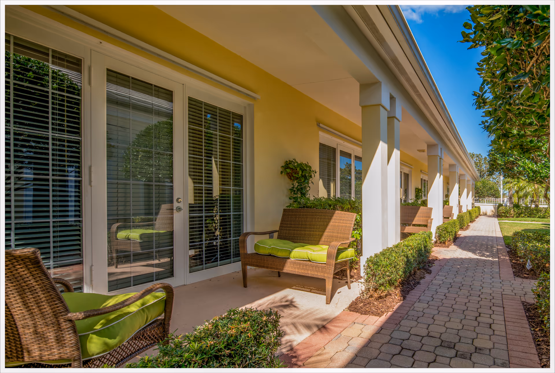 Covered patio outside a yellow building with wicker chairs with green cushions lining a paved walkway.